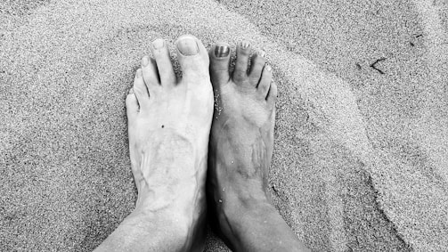 Two feet are placed side by side on a sandy surface. One foot is lighter in color and the other is darker. The skin texture and details of the feet are clearly visible. The sand around the feet appears fine and has subtle patterns created by the movement or placement of the feet.