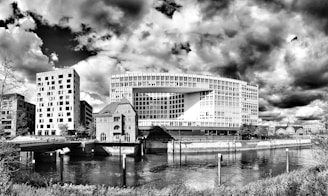 A striking architectural landscape features a modern office building with a unique rectangular design. Nearby, there are several other buildings, including one with a shorter, more traditional structure and a tower adorned with multiple windows. The scene is set against dramatic clouds, adding depth and texture to the black-and-white image. The foreground includes a calm river reflecting the buildings above, bordered by grassy embankments and shrubbery.