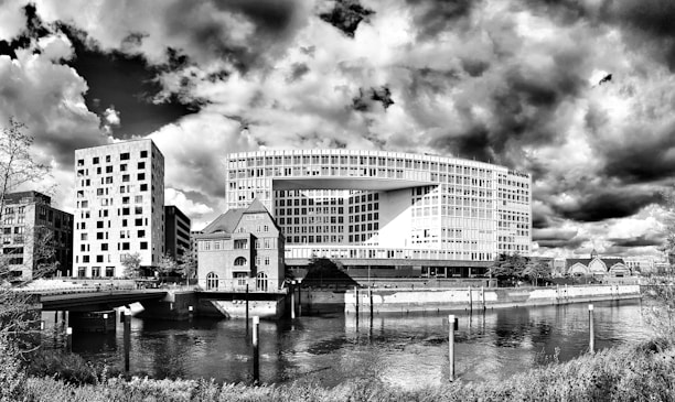 A striking architectural landscape features a modern office building with a unique rectangular design. Nearby, there are several other buildings, including one with a shorter, more traditional structure and a tower adorned with multiple windows. The scene is set against dramatic clouds, adding depth and texture to the black-and-white image. The foreground includes a calm river reflecting the buildings above, bordered by grassy embankments and shrubbery.