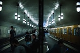 People are waiting and interacting with mobile devices at a dimly lit subway station, with a yellow train on one side and hanging lights from the ceiling.