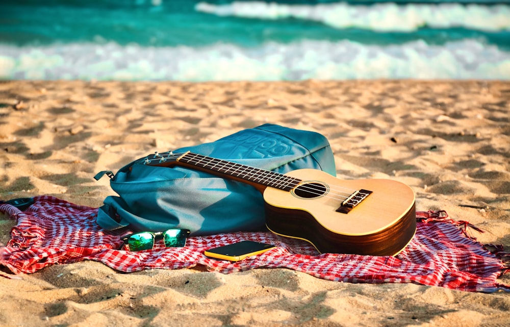 natural finish acoustic guitar near yellow tablet and sunglasses on red blanket placed on seashore