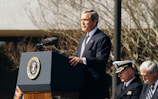 A man in a suit is standing at a podium delivering a speech. The podium has the seal of the President of the United States. To the right, there are two seated men, one in a military uniform and the other in a suit. Bare tree branches are visible in the background.