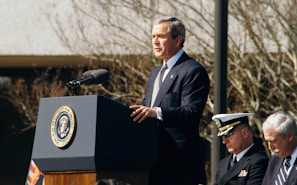 A man in a suit is standing at a podium delivering a speech. The podium has the seal of the President of the United States. To the right, there are two seated men, one in a military uniform and the other in a suit. Bare tree branches are visible in the background.
