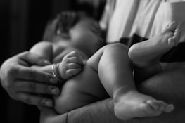 A close-up of a newborn baby sleeping peacefully in the arms of an adult. The focus is on the baby's tiny feet and hands, highlighting the gentle and protective grasp of the adult.