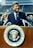 A man in a suit stands at a podium featuring the seal of the President of the United States. The setting appears to be an outdoor stadium with several people in the background, some of whom are applauding.