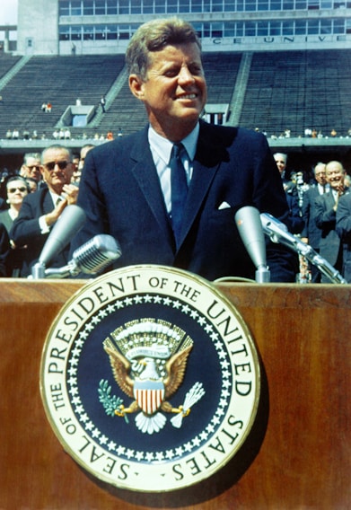 A man in a suit stands at a podium featuring the seal of the President of the United States. The setting appears to be an outdoor stadium with several people in the background, some of whom are applauding.