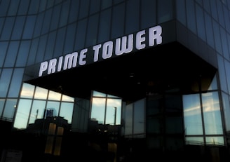 A modern building with a sleek glass facade reflecting the sky and surrounding structures, prominently displaying the name 'Prime Tower' in large, illuminated letters.