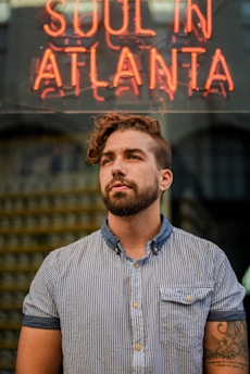 A person with a beard and tattoo stands in front of a neon sign displaying the words 'Soul in Atlanta.' The individual is wearing a striped button-up shirt and gazes upward, with a thoughtful expression. The background features a blurred reflection, adding depth to the scene.