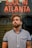A person with a beard and tattoo stands in front of a neon sign displaying the words 'Soul in Atlanta.' The individual is wearing a striped button-up shirt and gazes upward, with a thoughtful expression. The background features a blurred reflection, adding depth to the scene.