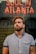A person with a beard and tattoo stands in front of a neon sign displaying the words 'Soul in Atlanta.' The individual is wearing a striped button-up shirt and gazes upward, with a thoughtful expression. The background features a blurred reflection, adding depth to the scene.