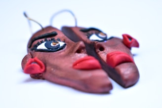 A close-up view of a unique earring made from clay that resembles a face, split into two pieces. The earring features painted eyes with dramatic eyeliner and bold red lips. There are metal hooks at the top for attaching to ears.