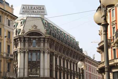A historic European building with ornate architectural details and a prominent sign reading 'Alleanza Assicurazioni' is situated on a busy street. The structure features decorative windows, column-like elements, and a roof adorned with elaborate designs. Surrounding buildings exhibit classic European architecture, with various colors and intricate facades. Streetlights and overhead wires can also be seen.