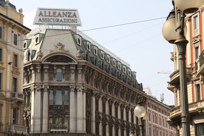 A historic European building with ornate architectural details and a prominent sign reading 'Alleanza Assicurazioni' is situated on a busy street. The structure features decorative windows, column-like elements, and a roof adorned with elaborate designs. Surrounding buildings exhibit classic European architecture, with various colors and intricate facades. Streetlights and overhead wires can also be seen.