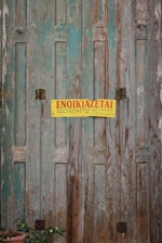 A weathered wooden door with peeling aquamarine paint, featuring a yellow sign with red text written in Greek. A terracotta pot with a small plant can be seen at the bottom left corner.