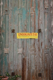 A weathered wooden door with peeling aquamarine paint, featuring a yellow sign with red text written in Greek. A terracotta pot with a small plant can be seen at the bottom left corner.