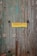 A weathered wooden door with peeling aquamarine paint, featuring a yellow sign with red text written in Greek. A terracotta pot with a small plant can be seen at the bottom left corner.