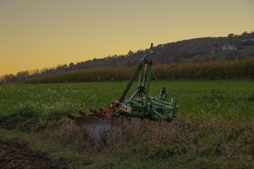 A piece of agricultural machinery, likely a plow, is positioned on lush green farmland during what appears to be early evening. The background shows rolling hills with patches of trees, and a clear sky transitioning from yellow near the horizon to a deeper shade above.