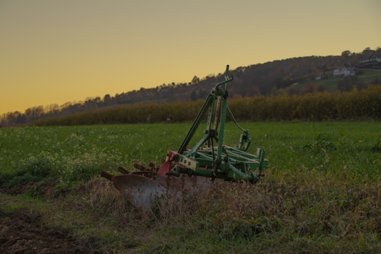 A piece of agricultural machinery, likely a plow, is positioned on lush green farmland during what appears to be early evening. The background shows rolling hills with patches of trees, and a clear sky transitioning from yellow near the horizon to a deeper shade above.