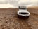A rugged white off-road vehicle is traveling on a rocky terrain under an overcast sky. The landscape is barren with scattered rocks and minimal vegetation, set against a backdrop of distant hills.