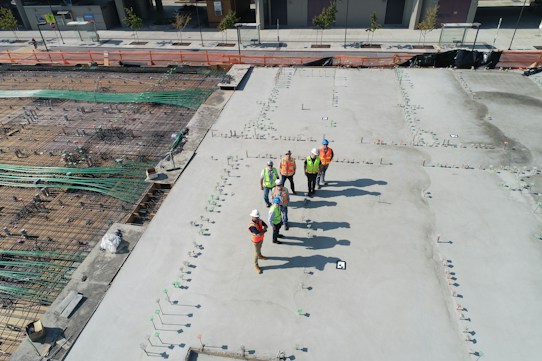A construction site with several workers wearing safety vests and helmets standing on a large concrete surface. The site contains visible steel reinforcements and construction materials. Urban landscaping and a city sidewalk can be seen in the background.
