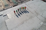 Six construction workers wearing safety vests and helmets stand on a large, recently poured concrete slab. The area is part of an active construction site with rebar grids visible in the background. The workers' shadows are long due to the angle of the sun.