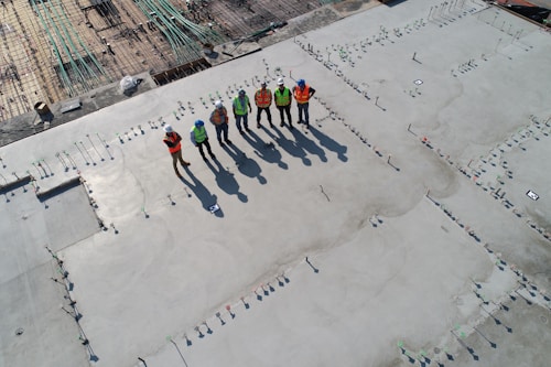 Six construction workers wearing safety vests and helmets stand on a large, recently poured concrete slab. The area is part of an active construction site with rebar grids visible in the background. The workers' shadows are long due to the angle of the sun.