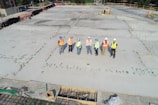 A group of construction workers wearing safety vests and hard hats stands on a large concrete construction site. The area is marked with stakes and rods, indicating future building work. The workers appear to be discussing or surveying the space. In the background, there are trees and a small structure under construction.