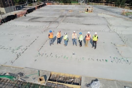 A group of construction workers wearing safety vests and hard hats stands on a large concrete construction site. The area is marked with stakes and rods, indicating future building work. The workers appear to be discussing or surveying the space. In the background, there are trees and a small structure under construction.