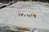A group of construction workers wearing safety vests and hard hats stands on a large concrete construction site. The area is marked with stakes and rods, indicating future building work. The workers appear to be discussing or surveying the space. In the background, there are trees and a small structure under construction.