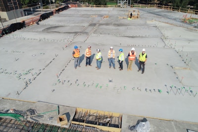 A group of construction workers wearing safety vests and hard hats stands on a large concrete construction site. The area is marked with stakes and rods, indicating future building work. The workers appear to be discussing or surveying the space. In the background, there are trees and a small structure under construction.