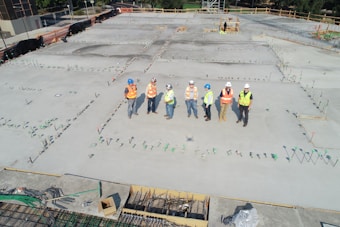A group of construction workers wearing safety vests and hard hats stands on a large concrete construction site. The area is marked with stakes and rods, indicating future building work. The workers appear to be discussing or surveying the space. In the background, there are trees and a small structure under construction.