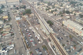 Aerial view of a large parking lot surrounding a train station, with various cars parked in neat rows. A train is visible on the elevated tracks running parallel to the parking area. Residential buildings and streets are in the background, creating an urban setting.