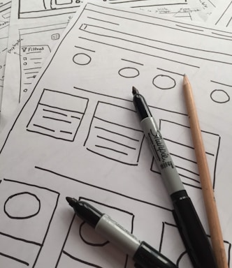 Several sheets of paper feature sketch-style website wireframes drawn with black marker lines. Two black markers and a wooden pencil rest on top of the papers, giving the scene a creative and informal touch.