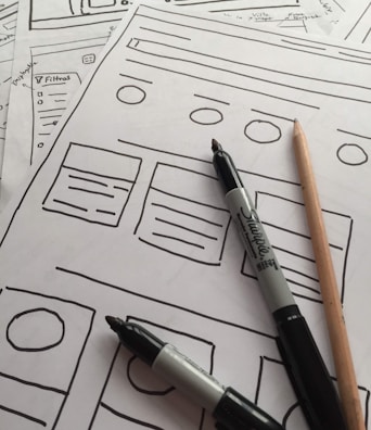 Several sheets of paper feature sketch-style website wireframes drawn with black marker lines. Two black markers and a wooden pencil rest on top of the papers, giving the scene a creative and informal touch.