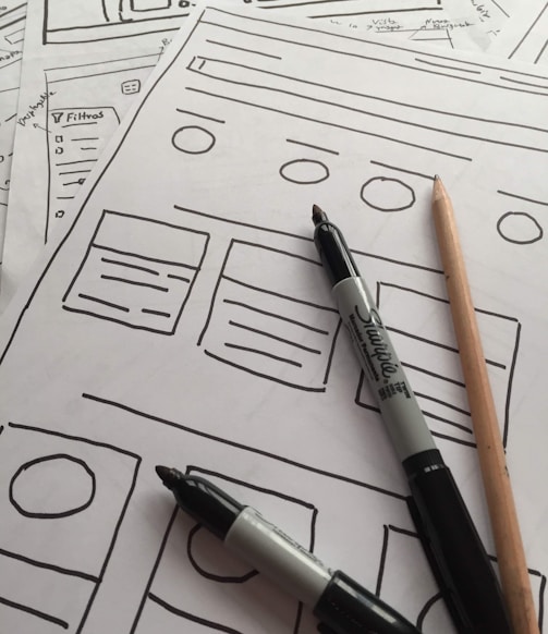 Several sheets of paper feature sketch-style website wireframes drawn with black marker lines. Two black markers and a wooden pencil rest on top of the papers, giving the scene a creative and informal touch.