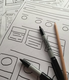 Several sheets of paper feature sketch-style website wireframes drawn with black marker lines. Two black markers and a wooden pencil rest on top of the papers, giving the scene a creative and informal touch.