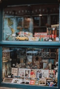 A shop window filled with various vintage and antique items, including a yellow toy car, maps, watches, photographs, and promotional signs. The display is organized with a world map as a backdrop, while model cars and memorabilia are arranged neatly in the foreground.