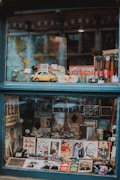 A shop window filled with various vintage and antique items, including a yellow toy car, maps, watches, photographs, and promotional signs. The display is organized with a world map as a backdrop, while model cars and memorabilia are arranged neatly in the foreground.