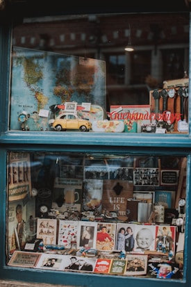 A shop window filled with various vintage and antique items, including a yellow toy car, maps, watches, photographs, and promotional signs. The display is organized with a world map as a backdrop, while model cars and memorabilia are arranged neatly in the foreground.