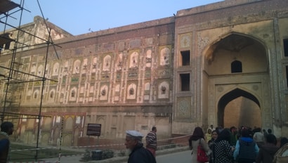 A large, intricately designed historic building features detailed stonework and numerous arched openings, showcasing traditional architectural elements. People are walking and gathering around the entrance, and there's scaffolding on the left indicating restoration work. The colors are muted and aged, suggesting an old structure.