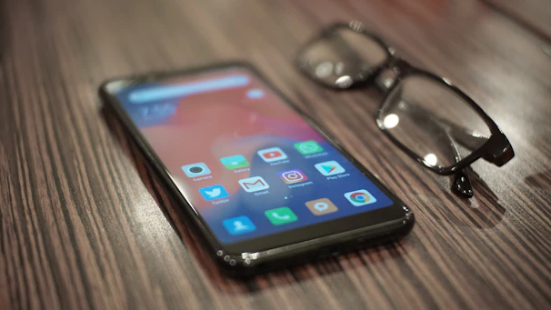 A smartphone resting on a wooden table next to a pair of glasses and a notebook.