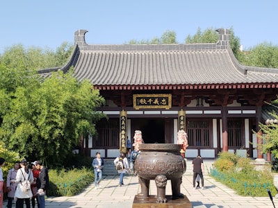 A traditional Asian temple with a tiled roof and ornate carvings, set amidst lush greenery. In front of the temple, there is a large, decorative bronze cauldron on a pedestal. A group of people are walking and conversing in the foreground.