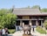 A traditional Asian temple with a tiled roof and ornate carvings, set amidst lush greenery. In front of the temple, there is a large, decorative bronze cauldron on a pedestal. A group of people are walking and conversing in the foreground.