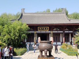 A traditional Asian temple with a tiled roof and ornate carvings, set amidst lush greenery. In front of the temple, there is a large, decorative bronze cauldron on a pedestal. A group of people are walking and conversing in the foreground.