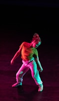 A dancer poses with a dynamic stance against a dark background, illuminated by colorful lights that cast hues of red and green. The dancer wears a bright yellow top and white pants, creating a vivid contrast with the darkness.