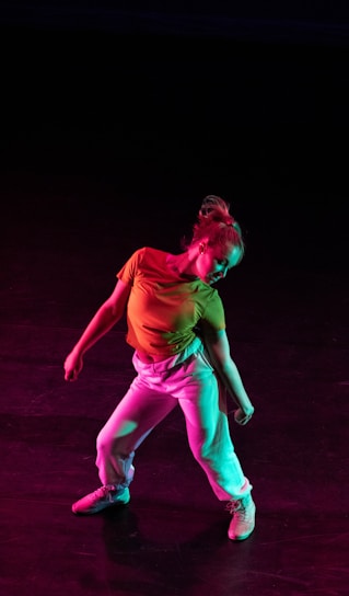 A dancer poses with a dynamic stance against a dark background, illuminated by colorful lights that cast hues of red and green. The dancer wears a bright yellow top and white pants, creating a vivid contrast with the darkness.