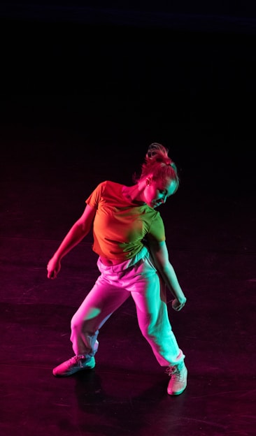 A dancer poses with a dynamic stance against a dark background, illuminated by colorful lights that cast hues of red and green. The dancer wears a bright yellow top and white pants, creating a vivid contrast with the darkness.