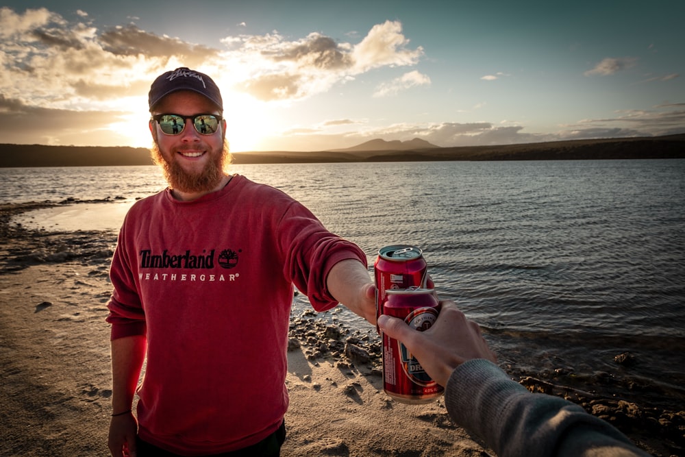 man in red sweater toasting red beverage can near body of water during daytime