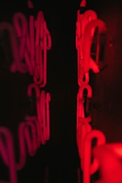 The image features red neon tubes arranged in a pattern within a dark setting, creating a glowing effect against the black background. The neon lights are reflected on a surface, enhancing the visual impact and adding depth.