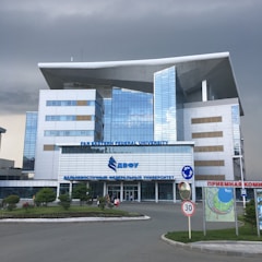 A modern building with a distinctive architectural design featuring large windows and a prominent overhanging roof. The facade is composed of reflective glass panels. It is identified as Far Eastern Federal University. Surrounding the building is a landscaped area with grass and small trees. There are some people near the entrance, and a 30 km/h speed limit sign along the road in front.